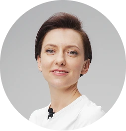 Kobieta z krótkimi, ciemnymi włosami, ubrana w białą bluzkę, która ma przypięty mikrofon do kołnierza, na jasnym tle. Zdjęcie w okrągłej ramce.