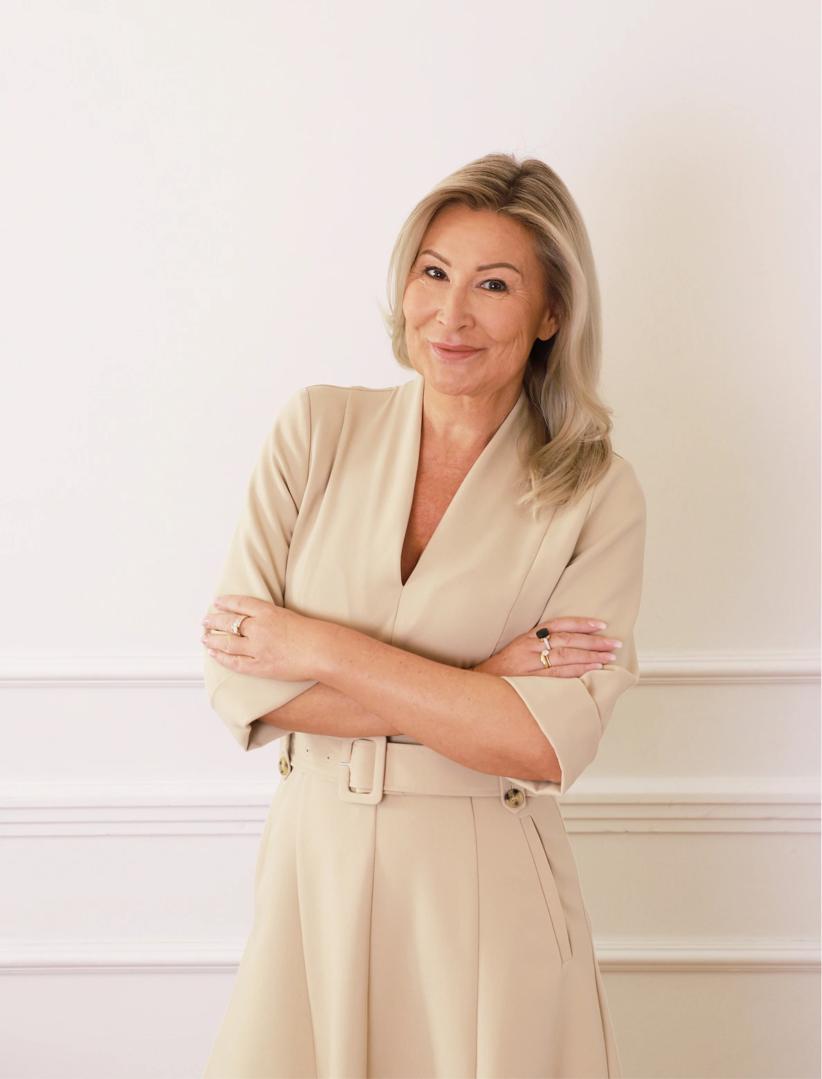 Kobieta o jasnych, falowanych włosach ubrana w beżową sukienkę z paskiem, stoi z założonymi rękami na tle jasnej ściany z dekoracyjnymi listwami.