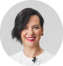 Kobieta z krótkimi, ciemnymi włosami zaczesanymi na bok, ubrana w białą bluzkę, która ma przypięty mikrofon do kołnierza, na jasnym tle. Zdjęcie w okrągłej ramce.